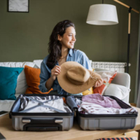 Young woman sitting on the couch and packing suitcase. She is holding a straw hat in her hands